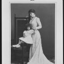 Victoria Melita, Grand Duchess of Hesse, stands looking down at her daughter, Princess Elizabeth, who is seated on the arm of a chair looking to front.